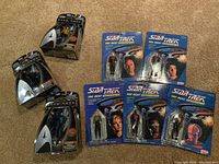 Eight Star Trek The Next Generation action figures displayed on carpet, some in silver emblem-shaped blister card packaging, others in blue card packaging.