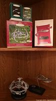 Four boxed and unboxed ornaments displayed on wooden shelf: two green-lidded boxes, one large red-lidded box, one white box, Deep Space Nine station on shelf and Enterprise NX-01 on lower shelf.