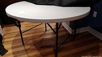 White round folding table with plastic top supported by black metal legs, standing on wooden floor