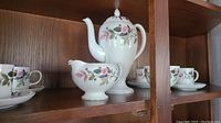 Photo showing coffee pot with lid, creamer, cups, and saucers arranged on shelf.