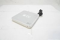 Top and angled view of the silver Apple TV 2007 media player with power cable visible, showing logos and surface wear.