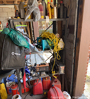 Image showing Motomaster and Terminator battery chargers on a metal shelf with hanging yellow rope and assorted cleaning fluid bottles above several red plastic gas cans on the floor.
