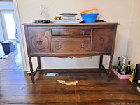 Front view of wood buffet with various household items placed on top, showing two drawers center and cabinet doors on sides, brass handles, turned legs.