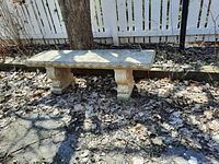 Side angle view of concrete garden bench placed outdoors on dirt and dried leaves next to tree and fence.