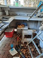 Photo showing two coolers (red/white and gray/blue), white step stool, orange and yellow extension cords tangled, blue outdoor glass on small white block, all outdoors with leaves and debris.