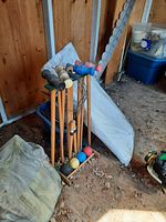 Wooden-handled croquet set with 6 mallets and 6 balls on a wooden rack, folded tent fabric leaning against the wall, plastic storage bin with lid nearby. Floor is covered with dirt and leaves.