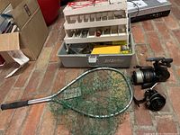 Open Lid Locker tackle box with multiple compartment trays filled with fishing tackle and accessories. Green fishing net and two fishing reels positioned in front of box.