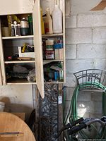 Shelves inside a white cabinet with assorted gardening supplies including bottles, spray cans, containers, and tools. A black metal lawn decoration stand with floral design leaning next to the cabinet.