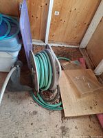 Plastic hose reel with green garden hose wound around it placed on dirt floor against wooden wall.