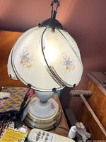 Table lamp with intact decorative white glass shade featuring floral designs and metal supports, ceramic base with floral painting, brass platform base. On a table with other household items.
