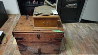 Photo of vintage Ideal 300 Columbia record player on top of wooden trunk, showing turntable, tone arm, and horn speaker arm with rustic wood and metal trunk below
