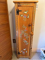 Front view of the tall wooden cabinet showing decorative hand painted designs of birds and floral vines on the door and top edges. Features black metal hinges and a wooden knob.