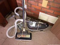 Wide shot of the upright vacuum cleaner with hose and attached power cord, showing the Turbo Brush head and metal handle, against a brick wall