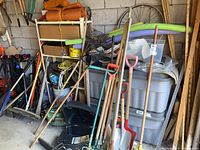 Corner view showing multiple long-handled tools leaning against wall, including rakes, shovels, axes, grass catcher bag and chainsaw box beneath.