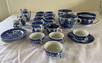 Front overview showing entire set of blue and white willowware ceramic tea set elements including creamer, sugar bowl, multiple tea cups, saucers, and plates arranged on cloth.