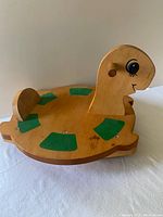 Top and side views of the wooden infant balance board shaped like a turtle, showing green grip pads and turtle face with wooden handles.