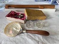 Photo showing fiddle bow bread slicer with cherry wood handle in background, abalone shell server with sterling silver collar and wooden handle in front, two vintage cut glass salt dishes, and silver tone bells in box.