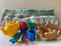 Plastic children's tea set with yellow tea pot, colorful cups, spoons, creamer, and plates, arranged with maple barn animal figures and a quilted blanket rolled up behind.
