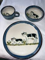 Set includes pottery plate, bowl, and cup with hand-painted cow illustrations and blue rim, arranged on white background