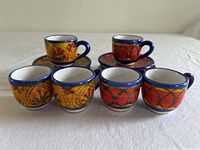 Six hand-painted espresso cups and matching saucers displayed on a table, showing their colorful artistic pattern and blue handles.