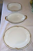 Three Limoges serving pieces displayed on a white cloth showing oval vegetable server, rectangular server with gold handles, and oval platter with floral and gold decorative trim.