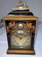 Front side view of the black and gold Tempus Fugit mantel clock with painted oriental decorations and a handle on top.