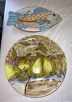 Two hand painted serving platters placed side by side on white surface, one pear design round plate and one fish shaped colorful plate
