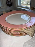Stack of eight pink tone plates with gold ring detailing around the inner edge, white center, on a white cloth surface.