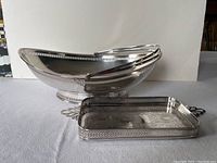 Two silver tone items displayed on a flat surface. Front item is a rectangular calling card tray with pierced sides and a decorative border. Back item is an oval serving centerpiece with a rounded pedestal base and monogrammed marking.