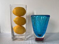 Clear rectangular glass vase with three large artificial lemons inside, beside hand-blown blue glass vase with rose-colored bottom on white cloth background.