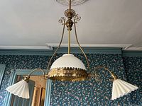 Full view of the chandelier showing the three glass shades and brass arms against floral wallpaper.