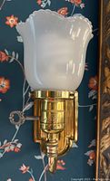 Close-up photo of a Victorian brass wall sconce showing the frosted white glass shade with scalloped edge and a wind-up key near the base.