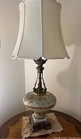 Full view of Victorian table lamp with white custom fabric lampshade and painted glass body on wooden table with lace doily.