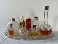 Full set of eight vintage perfume bottles and one talc bottle displayed on decorative cut glass tray. Various shapes and stopper styles with amber-colored contents visible.