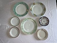 Seven assorted vintage plates arranged on a white tablecloth showing different sizes and floral decorative borders.