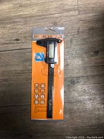 Front view of the digital caliper in sealed original packaging, showing black caliper with LCD display, included battery, and orange backing card.