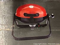 Photo of the portable propane grill inside the box showing the red powder-coated lid, black base, and attached handle.