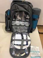 Picnic backpack opened showing cutlery in slots, 4 plastic wine glasses, plates, and a bottle opener in clear view.