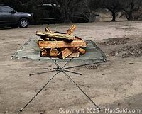 Firepit set up outdoors holding firewood, showing foldable tripod metal stand with stainless steel mesh surface.
