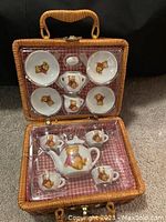 Wicker picnic basket open showing miniature porcelain tea set including teapot, four cups, and four plates with teddy bear designs.