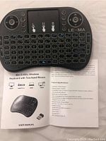 Photo showing front of wireless mini keyboard with integrated touchpad mouse, Lexma branding, and user manual with product specifications.