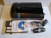 Photo showing Epson Perfection V500 scanner with accessories arranged below: film holders, software CD, power cable, USB cable, and printed manual in plastic wrap.