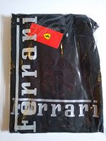 Front view of the sealed black T-shirt in plastic packaging showing Ferrari logo and red Ferrari tag.