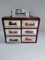 Wooden display case with seven die cast antique cars in original boxes arranged in compartments, top view.