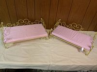Two vintage metal doll beds with pink mattresses set on a white surface against wood panel background.
