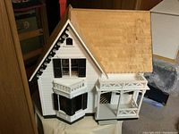 Front side view of the wooden Victorian-style dollhouse showing textured wooden shingle roof, white siding, black shutters, and front porch with decorative railings.