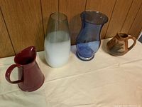Four jugs and vases displayed on a white surface against wood panel background, including two glass vases and two ceramic jugs.