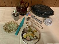 Full view showing the ceramic tea pot, roast pan with lid, towel rack, spoons, birthday candle holders, decorative plate, knife, and ice cream scoop.