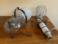 Overview of three lights on wooden table: large glass hanging candle holder with metal band and chain, battery-powered light device with wires, small cage-framed electric bulb.