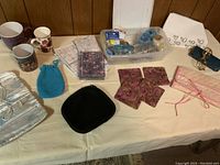 Wide view of the miscellaneous lot items arranged on a table including mugs, coasters, trays, pouches, and plastic container with small items.